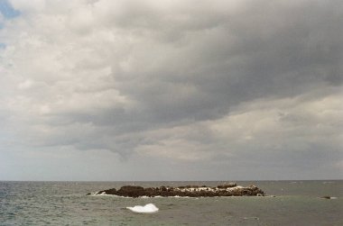 Şiddetli fırtına bulutları gri bir deniz ve karanlık kayalık bir adanın üzerinde toplanırken dalgalar ön planda yavaşça kırılarak karamsar ve dramatik bir deniz manzarası oluşturur..