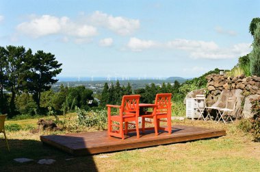 Parlak kırmızı sandalyeler ve küçük bir masa, yeşil bir kırsal manzaraya bakan ahşap bir güvertede oturur. Mavi gökyüzünün altında, yumuşak bulutlu uzak rüzgar türbinleri vardır..