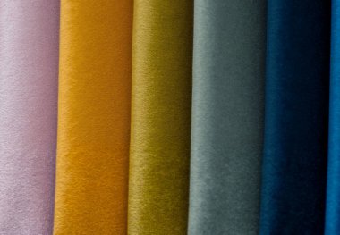 Kadife kumaş örnekleri dikey sıralanmış, zengin bir renk paleti sergiliyorlar. Soldan sağa doğru, renkler leylak, hardal, zeytin, gri, deniz mavisi ve koyu mavi tonlarını içerir. Kumaşlar yumuşak bir doku ve lüks bir parıltı sergiliyor..