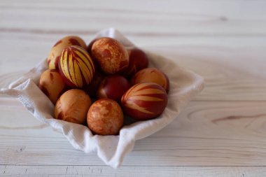 Paskalya yumurtalarıyla dolu bir sepet, açık ahşap bir masaya yerleştirilmiş. Yumurtalar çeşitli desenlerle kahverengi, turuncu ve kırmızıya boyanmıştır. Sepet beyaz bir bezle kaplanmış, yumurtaları güvenli bir şekilde tutuyor..