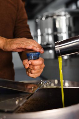 Sıcak tonlarda zeytin değirmeninin üzerinde mavi bir bardağı tutan bir el vurgulanıyor, burada taze saf zeytinyağı akıyor. Ayarlar, tatma sürecinin kaliteyi güvence altına aldığı geleneksel bir zanaatkar tesisini gösteriyor..