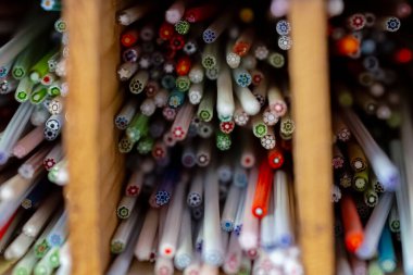 Ahşap bir kutuda Murrine cam bastonlar canlı renkler ve karmaşık geometrik desenler sergiliyor. Her çubuk, uçlarında küçük çiçek benzeri motiflerle eşsiz bir millefiori tasarımı içeriyor. Bu dekoratif çubuklar İtalya, Murano 'da yapıldı.