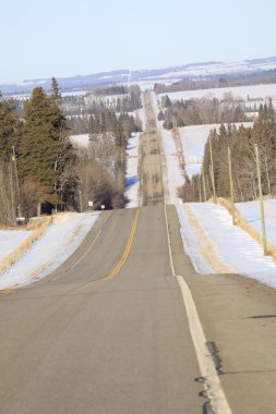Bu görüntü, kışın yuvarlanan bir arazide uzanan kırsal bir karayolunu ya da kır yolunu yakalar. Perspektif, ülkenin topoğrafyasından sonra yolun 
