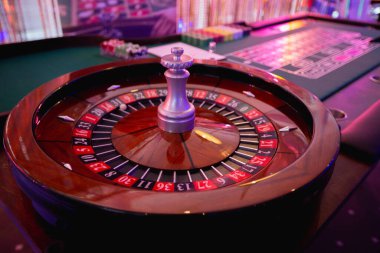 Lüks bir kumarhane rulet masasına yakın plan, renkli neon ışıklarla aydınlatılmış top hareket halinde. Zarif ahşap masa ve klasik kırmızı ve siyah numaralı düzen canlı bir kumar ortamı yaratıyor..