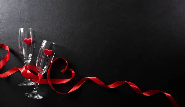 Valentine\'s day and love concept made from champagne glasses and red hearts on black wooden background. Top view with copy space, flat lay.