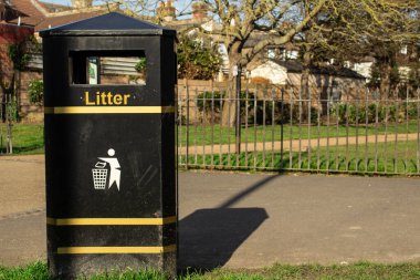 Parkta sarı çizgili siyah bir çöp kutusu. Arka planda demir bir çit ve bir yol var..
