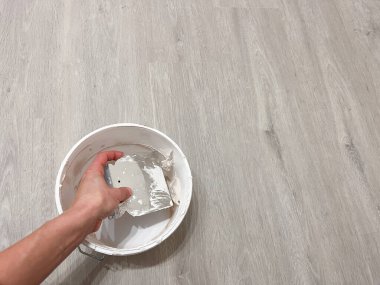 Tahta zemine yerleştirilmiş bir kovadan macunu çıkarmak için spatula kullanan bir elin yakın çekimi duvar onarımı ve yenileme çalışmalarını gösteriyor..