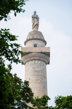 Bir adamın heykeliyle süslenmiş görkemli bir sütun, yemyeşil ağaçların çerçevelediği bulutlu bir gökyüzüne çarpar..