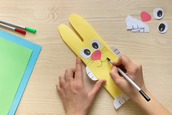 do it yourself: Easter sweets decor: wrapping a chocolate bar in the shape of an Easter bunny. Step 13: human hands draw the face, mustache, mouth to the Easter bunny, next to a light wooden background, colored paper and pens, a chocolate bar