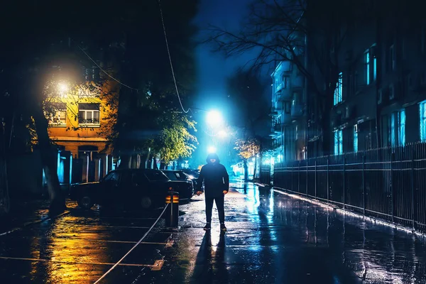 Silhouette of alone stranger in hood at night city street in rain. Creepy killer or stalker, criminal stands in shadow with urban lights reflected in puddles. Thriller horror mysterious atmosphere