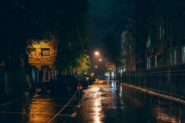 Empty city street in rainy weather at night illuminated by city lamps, no people, wet and puddles with reflection, horror and mystery atmosphere