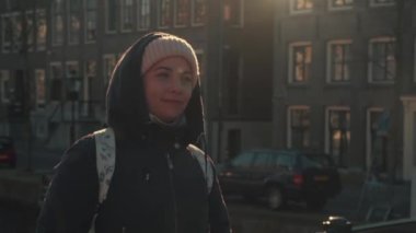 Amsterdam 'da bahar güneşli bir günde eski dans evlerinin arka planında şapkalı ve başlıklı sevimli bir gezgin kız. Avrupa seyahati kavramı.
