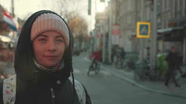 Portrait of cute girl in hat and hood with backpack on blurred background of street in historic center of Amsterdam — Stok Video