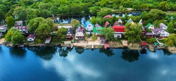 Small suburban houses on river bank with blue sky reflection in water and green trees forest on background, aerial view from drone