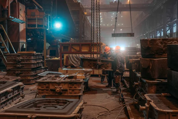 Los trabajadores en cascos y máscaras faciales trabaja con molde en cadenas de grúa después de la fundición de metal en la fundición de la fábrica de acero — Foto de Stock