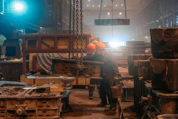 Pekerja dalam helm dan topeng wajah bekerja dengan cetakan pada rantai derek setelah pengecoran logam di pengecoran pabrik baja pengecoran — Stok Foto
