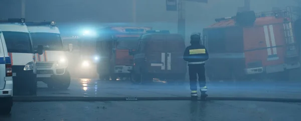 Birçok ambulans, itfaiye kamyonu, kurtarma araçları ve sağlık görevlilerinin bulunduğu yangın nedeniyle kapalı sokak panoraması duman altında. Kurtarıcı merkezde duruyor, arkadan bakıyor. Kazalar ve felaketler — Stok fotoğraf