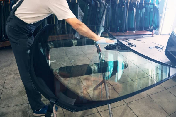 Automobile glazier worker degreases glass windscreen or windshield before installation on car in service station garage Stok Lukisan  