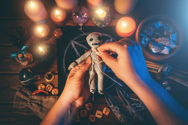 Sorceress or witch sticks needles into voodoo doll at ritual table with pentagram, burning candles and other occult objects, top view. Voodoo witchcraft, spirituality and occultism concept