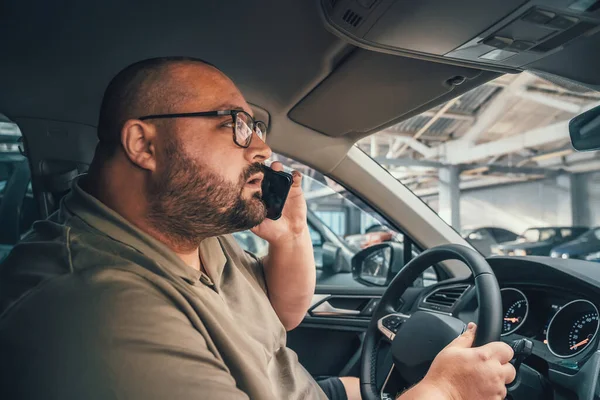 Pria berkacamata berbicara di telepon genggam saat mengendarai mobil di tempat parkir. Stok Gambar Bebas Royalti