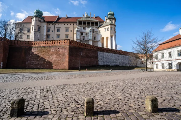 Polonya 'nın tarihi bir sembolü olan görkemli Wawel Kraliyet Kalesi' nin alçak açılı görüntüsü. Görüntü, heybetli kırmızı tuğla ve taş savunma duvarı, yeşil kubbeli Rönesans kuleleri ve öndeki meydanın kaldırımını vurguluyor.