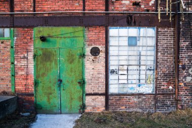 Polonya 'nın Gdansk kentindeki İmparatorluk Tersanesi' ndeki (Stocznia Cesarska) tarihi kırmızı tuğlalı sanayi duvarının yakın plan çekimi. Sahnede paslı yıpranmış yeşil metal bir kapı, grafiti etiketleriyle kaplı büyük bir sanayi penceresi ve
