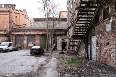 Polonya 'nın Gdansk kentindeki İmparatorluk Tersanesi' ndeki (Stocznia Cesarska) tarihi tuğla binaların dış görünüşü. Çekimde park halindeki arabalı bir avlu, duvar yazısıyla kaplı bir cephe boyunca paslı bir metal yangın merdiveni ve büyük terk edilmiş bir sanayi yer alıyor.