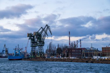 Gdansk 'taki Stocznia Cesarska' nın (İmparatorluk Tersanesi) panoramik manzarası, iskele üzerinde yükselen ikonik yeşil gantry vinçlerini içeriyor. Görüntü, limanın endüstriyel mirasını, tarihi tuğla binalar, tüten bacalar ve bir Ziyaretçi ile yakalıyor.