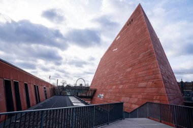 Gdansk 'taki İkinci Dünya Savaşı Müzesi' nin eğimli kırmızı beton kulesine bir bakış açısı. Görüntü, tarihsel notları sembolize eden, bulutlu bir gökyüzüne karşı inşa edilen kalın geometrik tasarımı ve benzersiz kısa piramit şeklini yakalar.