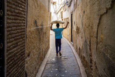 Fes el-Bali Medina 'nın dar labirentinde yürüyen Faslı bir çocuğun çarpıcı bir arka görüntüsü, UNESCO Dünya Mirası sahası. Genç adam ustalıkla kafasında geleneksel bir tepsi ekmek taşıyor, antik dokulu duvarlar çerçeveli ve...