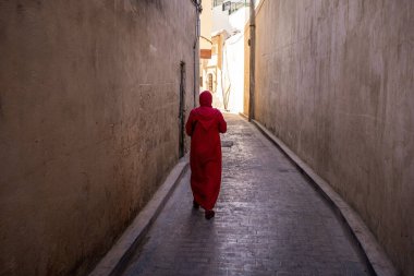 Tarihi Kazablanka 'nın Medine bölgesinde dar bir sokakta yürürken geleneksel kırmızı djellaba giyen bir kadının çarpıcı bir arka görüntüsü. Yüksek taş duvarlar ve gölgeler ve güneş ışığı arasındaki zıtlık zamansız bir atmosfer yaratır, Typi.