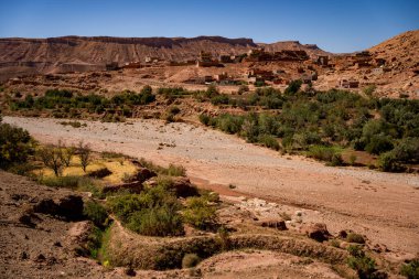 Çarpıcı bir panoramik manzara, bir çöl yamacına tünemiş eski bir Berber köyünü (ksar) yakalıyor. Görüntü, gür yeşil bitki örtüsüyle benekli kuru bir nehir yatağı (oued) ile engebeli kayalık arasında keskin bir kontrastı vurgular.