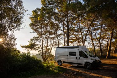 Altın saat boyunca uzun çam ağaçlarının arasına park etmiş modern beyaz bir karavanın çarpıcı görüntüsü. Sıcak günbatımı ışığı dalların arasından süzülerek doğanın göbeğinde özgürlük, macera ve vahşi bir kamp ortamı yaratır..