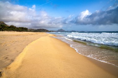 Güneydoğu Sardunya, İtalya 'daki Costa Rei sahilinin manzaralı bir sahili. Görüntü, ufukta bir Dyna 'nın altında dağlık bir siluet tarafından çerçevelenmiş karakteristik altın kum ve dalgalarıyla geniş, vahşi kıyı şeridini sergiliyor.