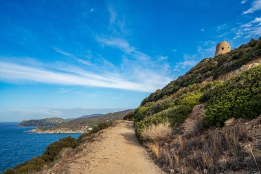Sinnai yakınlarındaki Sardinya 'nın güney kıyısında kıvrılan toprak bir patikanın panoramik görüntüsü. Görüntü karakteristik Akdeniz çalılığı ve denize bakan tarihi 16. yüzyıl Capo Boi kıyı kulesi, berrak mavi sular ve di