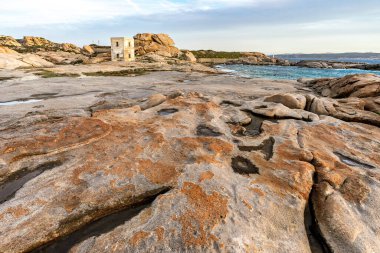 Punta Tegge 'nin La Maddalena adasındaki panoramik manzarası. Görüntü, Maddalena Takımadaları Ulusal Parkı 'nın karakteristik bir özelliği olan rüzgar heykeli şeklindeki engebeli bir kıyı manzarasına karşı kurulmuş antik bir taş gözlem kulesini gösteriyor..