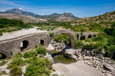 Güney Arnavutluk 'taki kristal berrak Mavi Göz pınarının yüksek açılı görüntüsü. Görüntü, Bistric nehrinin canlı turkuaz ve derin mavi sularını sergiliyor. Dalları sarkık ağaçlar ve sakin doğal ortamdaki yeşil su bitkileri tarafından çerçevelenmiş.