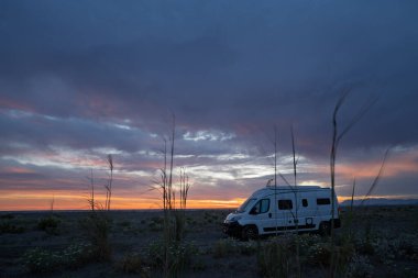 Dönüşmüş beyaz bir karavan altın saat boyunca vahşi bir kıyı manzarasında duruyor. Gökyüzünde dramatik bulutlar ve turuncu renkler, ön planda yerli kum tepecikleri bitki örtüsü bulunur. Özgürlük, vanlife ve yolculuk kavramları için mükemmel bir imaj.