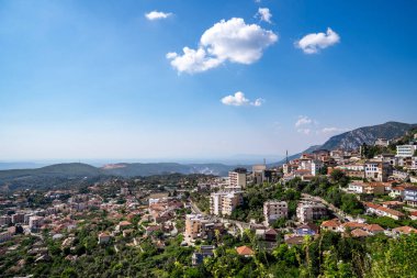 Kruja 'nın dağ sırtları arasına yerleştirilmiş kentsel manzarasını gösteren yüksek açılı bir perspektif. Görüntü, geleneksel evler, modern binalar ve açık mavi gökyüzü altında dağınık bulutlar altında yemyeşil alanlar arasındaki zıtlığı vurguluyor. Şeyde...