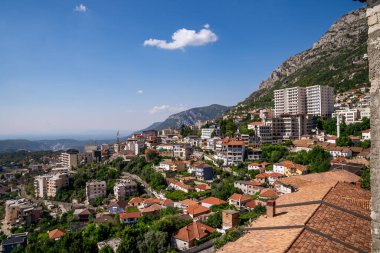 Kruja 'nın dağ sırtları arasına yerleştirilmiş kentsel manzarasını gösteren yüksek açılı bir perspektif. Görüntü, geleneksel evler, modern binalar ve açık mavi gökyüzü altında dağınık bulutlar altında yemyeşil alanlar arasındaki zıtlığı vurguluyor. Şeyde...