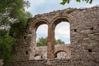M.S. 6. yüzyılda UNESCO 'nun Dünya Mirası adlı Butrint arkeoloji sahasında yer alan Büyük Bazilika' nın tuğla ve taş kemer ayrıntılarını gösteren yakın plan bir görüntü. Erken dönem Hıristiyan yapısı yemyeşil Akdeniz sebzeleriyle çevrilidir.