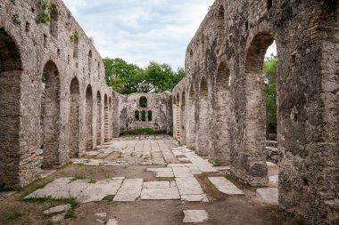 UNESCO 'ya ait bir Dünya Mirası olan Butrint Arkeolojik Alanı içindeki Büyük Bazilika' nın göbeğini gösteren bir perspektif. Görüntü, iyi korunmuş taş kemerleri ve uzaktaki maymunu vurguluyor. Natio 'nun yemyeşil yeşilliğine karşı kurulmuş.