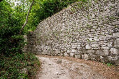 UNESCO 'nun Dünya Mirası sitesi Butrint' in devasa taş kent duvarlarının zemin katmanlı görüntüsü. Tahkimatlar, Yunan ve Roma 'nın yüzyıllarını gözler önüne seren arkeoloji parkının bereketli Akdeniz ormanları içinde toprak bir yolun yanında yer alıyor.