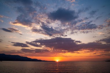 Arnavutluk 'un Ksamil kıyısından yakalanan İyon Denizi' nin ufkun altında batan güneşin nefes kesici manzarası. Görüntü, kıyı dağlarının siluetleriyle altın ışığı filtreleyen dağınık bulutlu canlı bir gökyüzünü ve