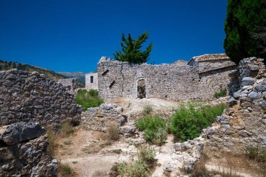 Arnavutluk Rivierası 'ndaki eski Himara Şatosu köyü içindeki tarihi yıkıntıların ve dar yolların zemin katındaki manzarası. Fotoğraf, Akdeniz yeşillikleriyle çevrili yerel taş duvarların yıkık dökük güzelliğini gösteriyor.