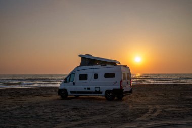 Adriyatik Denizi 'nin kumlu sahiline canlı bir günbatımında parketmiş beyaz bir karavan görüntüsü. Görüntü, vanlife hareketinin özgürlük ve macera duygusunu vurguluyor.
