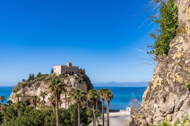 Tanrıların Sahili 'nin sembolü olan Tropea' daki Santa Maria dell 'Isola' nın ikonik Mabedi 'nin panoramik görüntüsü. Antik taş kilise, turkuaz deniz ve Akdeniz palmiyeleriyle çevrili yüksek bir uçurumun üzerinde, berrak mavi bir ağacın altında görkemli bir şekilde duruyor.