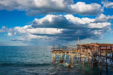 Rocca San Giovanni, Abruzzo 'daki Sasso della Cajana trabocco' nun panoramik görüntüsü. Bu geleneksel ahşap balıkçı makinesi Adriyatik Denizi 'nde ortaya çıkan hassas bir gökkuşağının yer aldığı büyük beyaz bulutlarla dolu mavi bir gökyüzüne karşı duruyor. Gerçek...