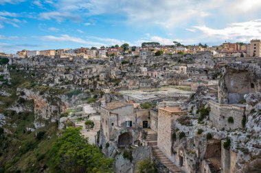 Matera 'nın tarihi merkezine geniş açılı muhteşem bir bakış açısı. Görüntü, Sasso Mağara Bölgesi 'nin antik mağara evlerini ve taş binalarını vurguluyor. Dağınık bulutlu mavi bir gökyüzünün altındaki yabani Gravina geçidine bakıyor..