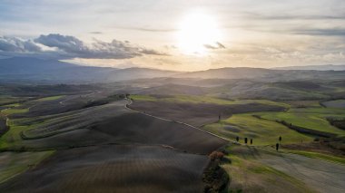 An evocative drone panorama showing the rolling hills of the Val d'Orcia at sunset. The warm sunlight illuminates the plowed fields and green meadows, highlighting the natural textures of the farmland and characteristic solitary cypress trees in the